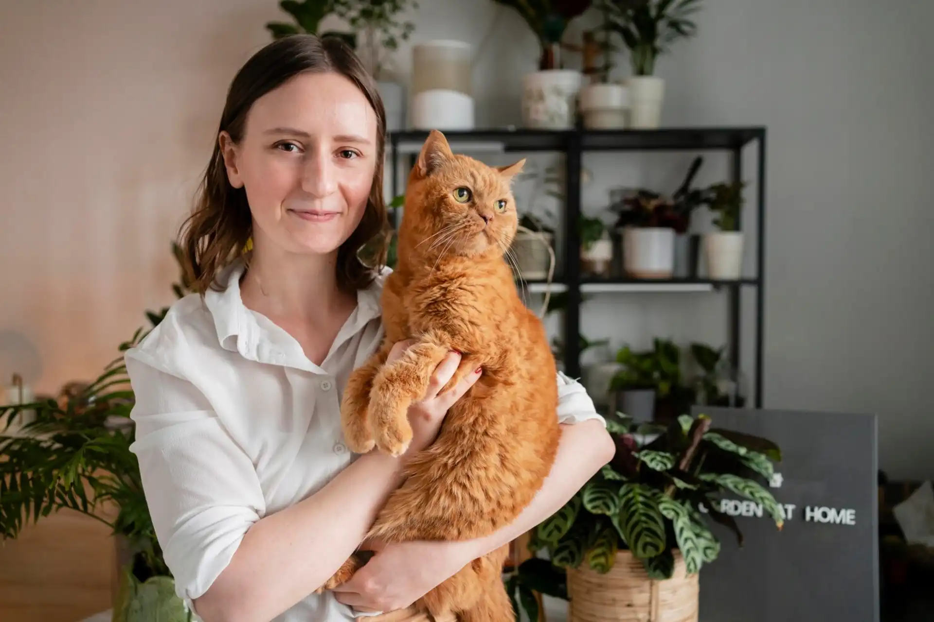Woman holding a ginger cat in a plant-filled home